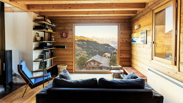 L’intérieur d’un chalet en bois chaleureux et accueillant, avec une grande fenêtre donnant sur les montagnes, une bibliothèque, un canapé et une petite table.