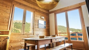 Une cabane en bois dotée de grandes fenêtres, d’une table à manger et d’un luminaire suspendu.
