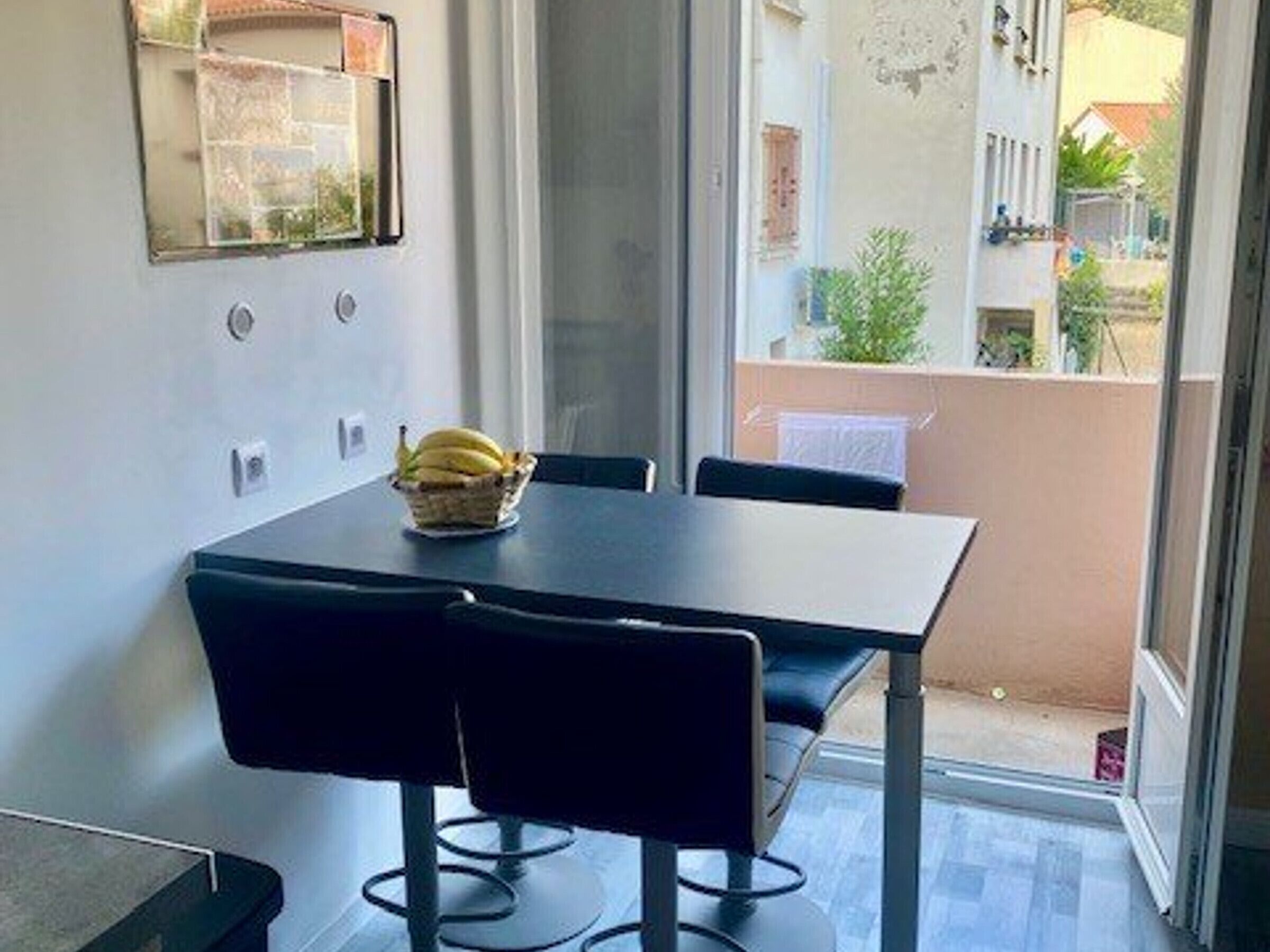 A dining area with a table and chairs, a basket of bananas on the table, and a view of a balcony with plants and a building in the background.
