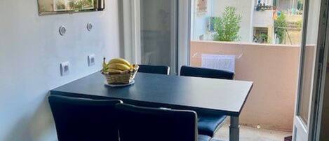 A dining area with a table and chairs, a basket of bananas on the table, and a view of a balcony with plants and a building in the background.