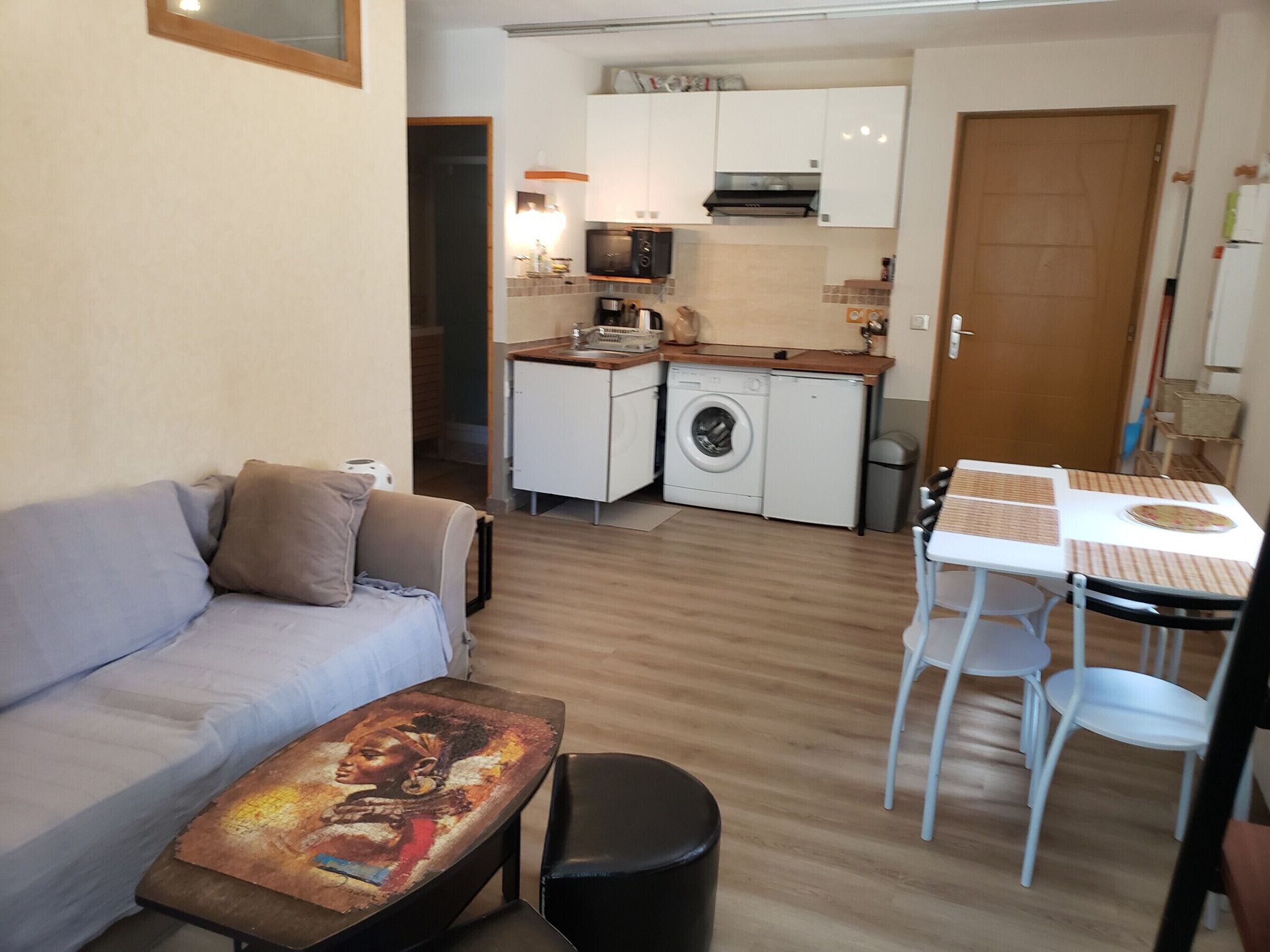Een compacte leefruimte met een bank, een klein tafeltje met tafelmat, een wasmachine en een kitchenette.