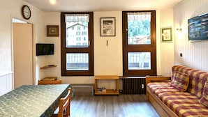 Ein gemütliches Zimmer mit Holzboden, einem grün gemusterten Bett, einer Couch mit kariertem Überwurf, einem Fernseher an der Wand und Fenstern mit Vorhängen.