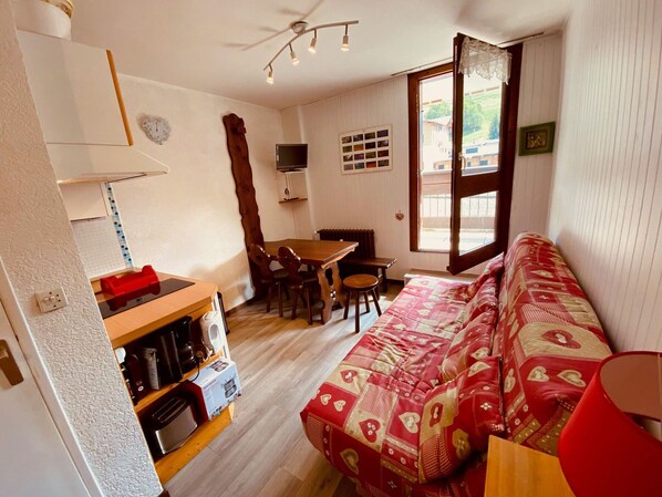 Ein gemütliches Wohnzimmer mit einer Couch, einem Esstisch und Stühlen. Es gibt eine Küchenzeile mit Geräten und ein Fenster mit Blick nach draußen.