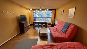 A compact living room with a red sofa, a television on a wooden cabinet, a dining table with chairs, and a window with curtains.