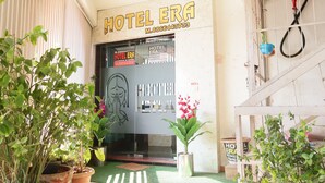 L’entrée d’un hôtel, avec une enseigne indiquant « HÔTEL ERA » et un numéro de téléphone. On y voit des plantes en pot et une sonnette fixée au mur.