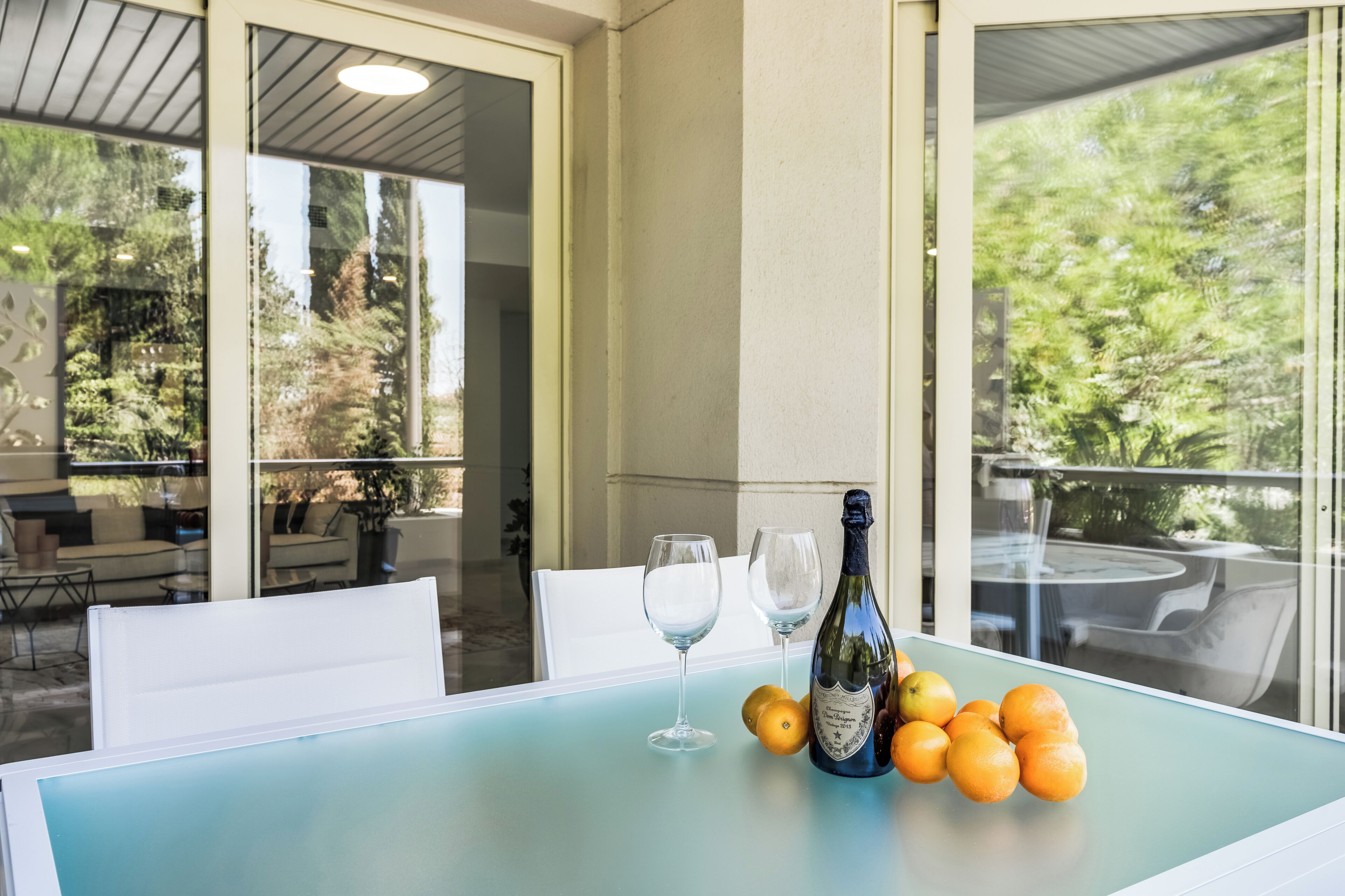 A table with a bottle of champagne, two glasses, and several oranges on a light blue table in a sunlit outdoor area with glass walls and furniture.