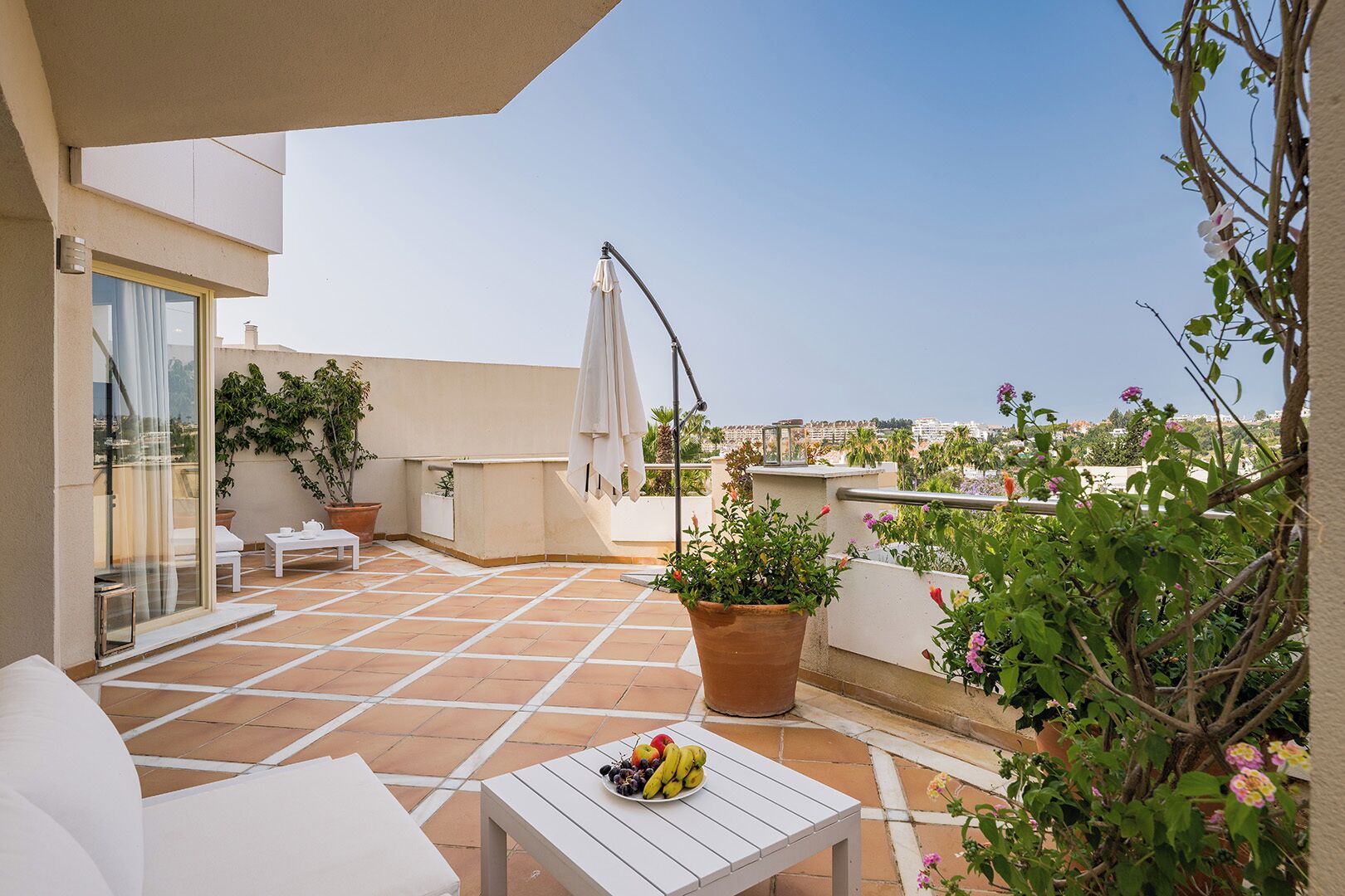 Una terraza en la azotea con mobiliario blanco, una planta en maceta y un frutero.