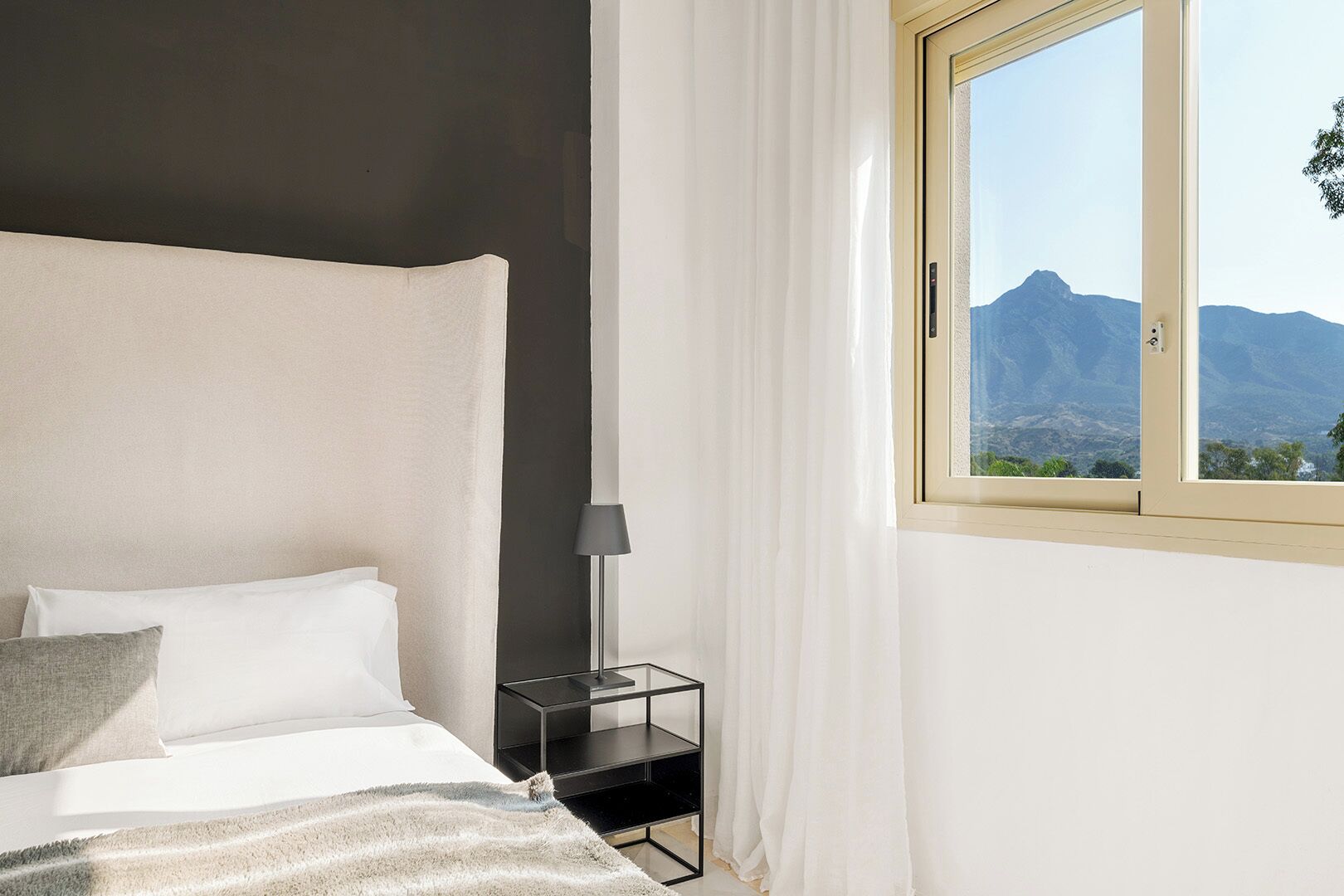 A bedroom with a bed, a nightstand, a lamp, and a window with a mountain view.