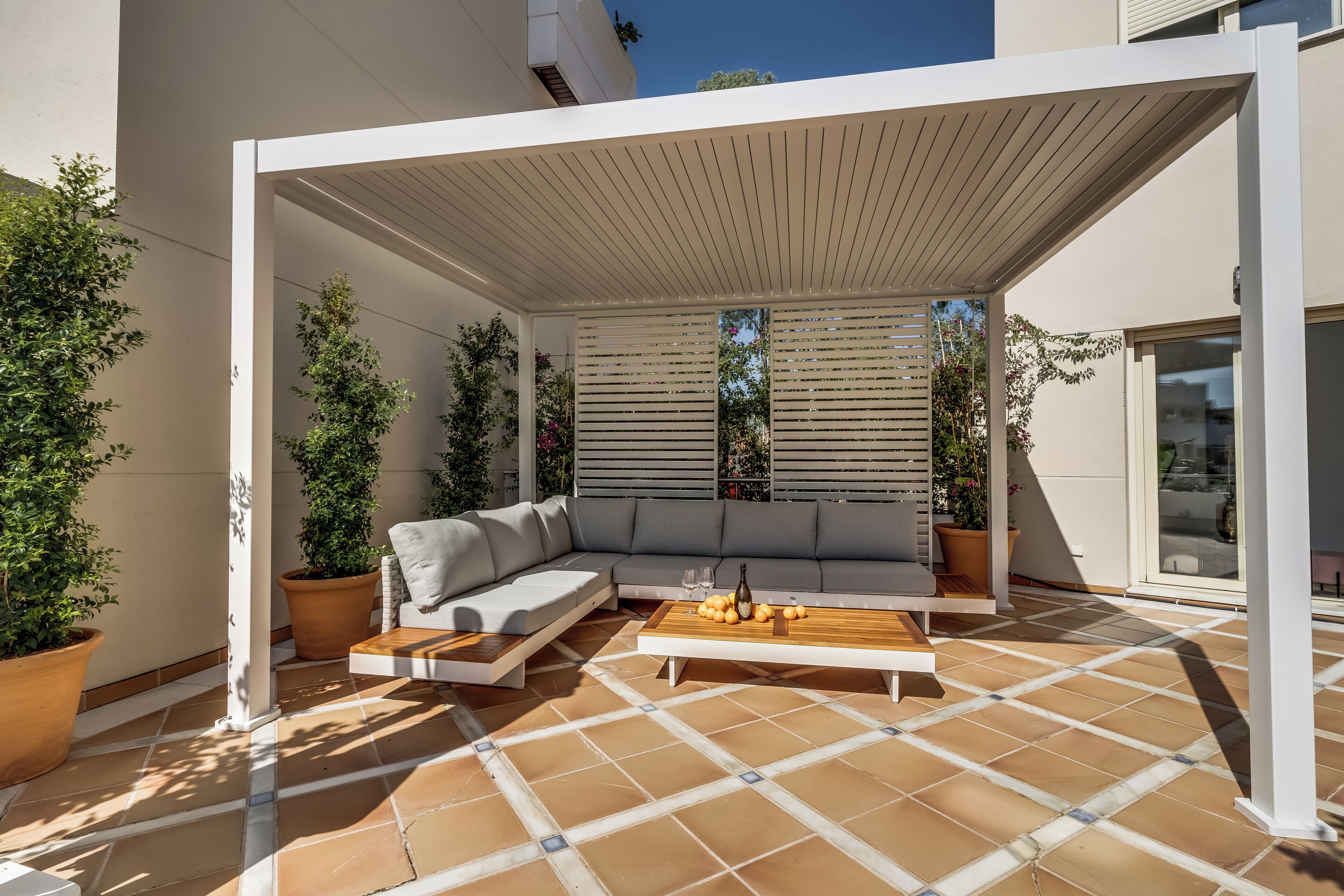 Un patio exterior moderno con una pérgola blanca, un sofá gris y una mesa de centro de madera.