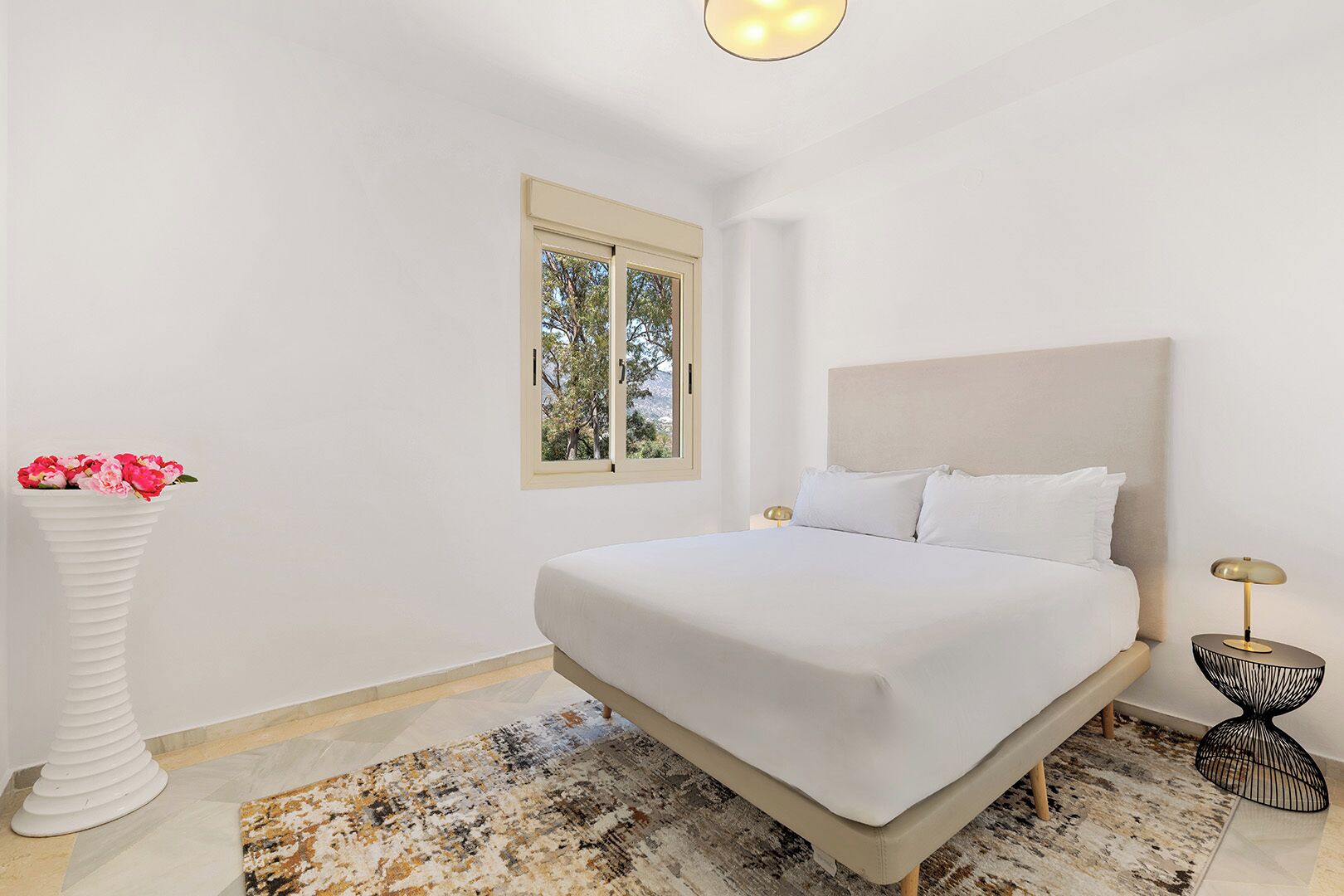 Een slaapkamer met een bed, een raam, een vaas met bloemen en een vloerkleed.