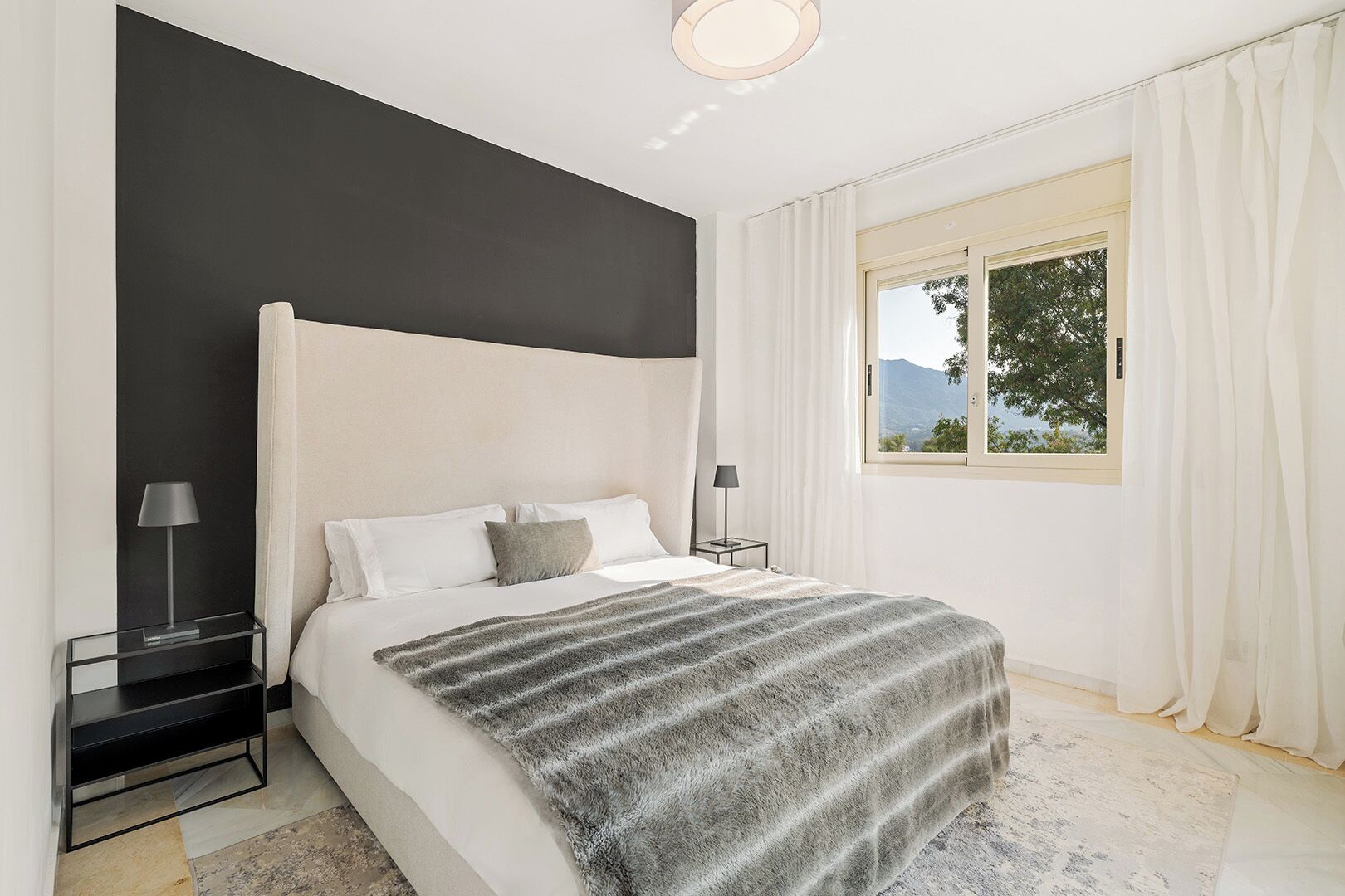 A modern bedroom with a large bed, two bedside tables, a view of the outdoors, and a black accent wall.