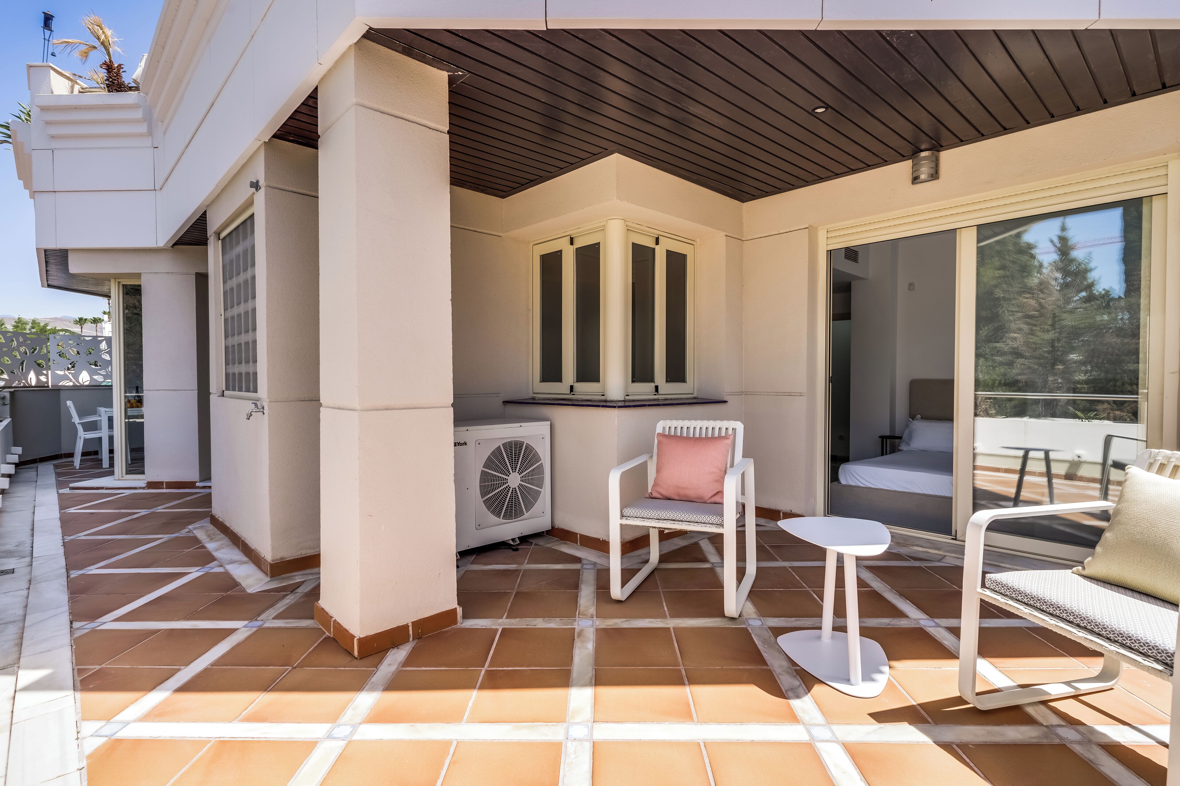 Une terrasse extérieure couverte, dotée d’un sol carrelé, d’une chaise blanche, d’une petite table et d’un canapé.