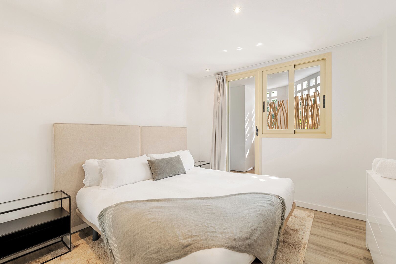 A modern bedroom with a large bed, a bedside table, and a window with a view of the outdoors.