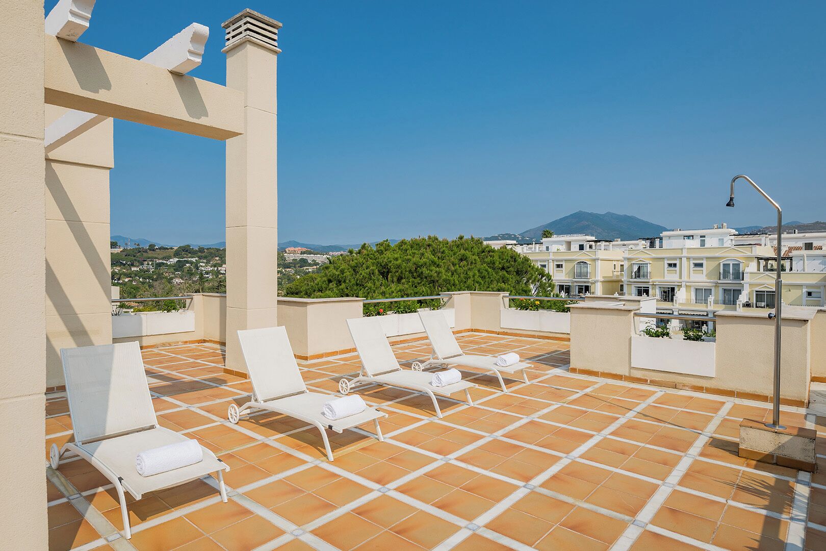 A rooftop terrace with white lounge chairs and a tiled floor, overlooking a residential area with buildings and greenery.