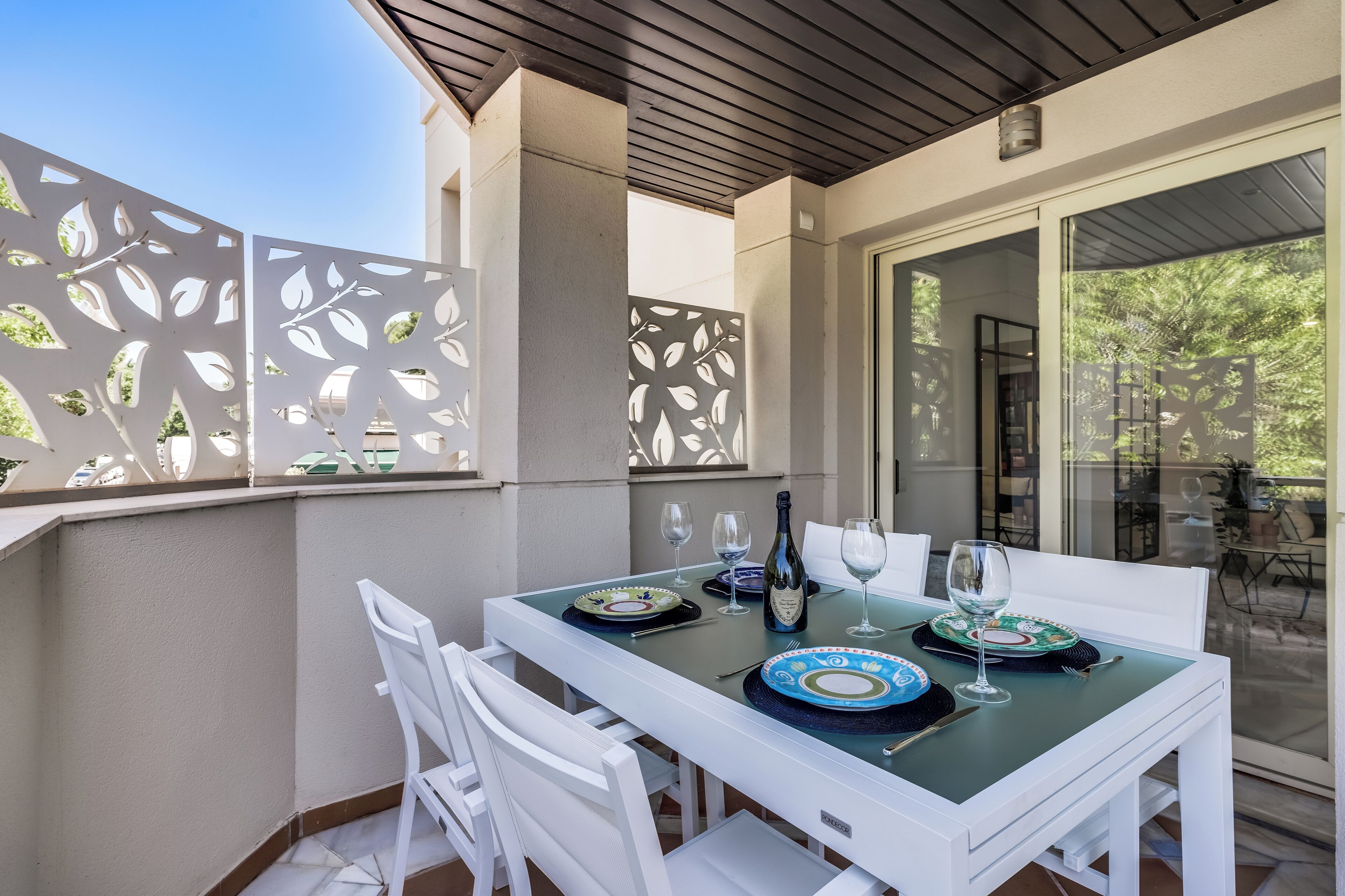 Un espacio de comedor al aire libre con una mesa dispuesta para dos, sillas blancas y un separador de vidrio con motivos de hojas.