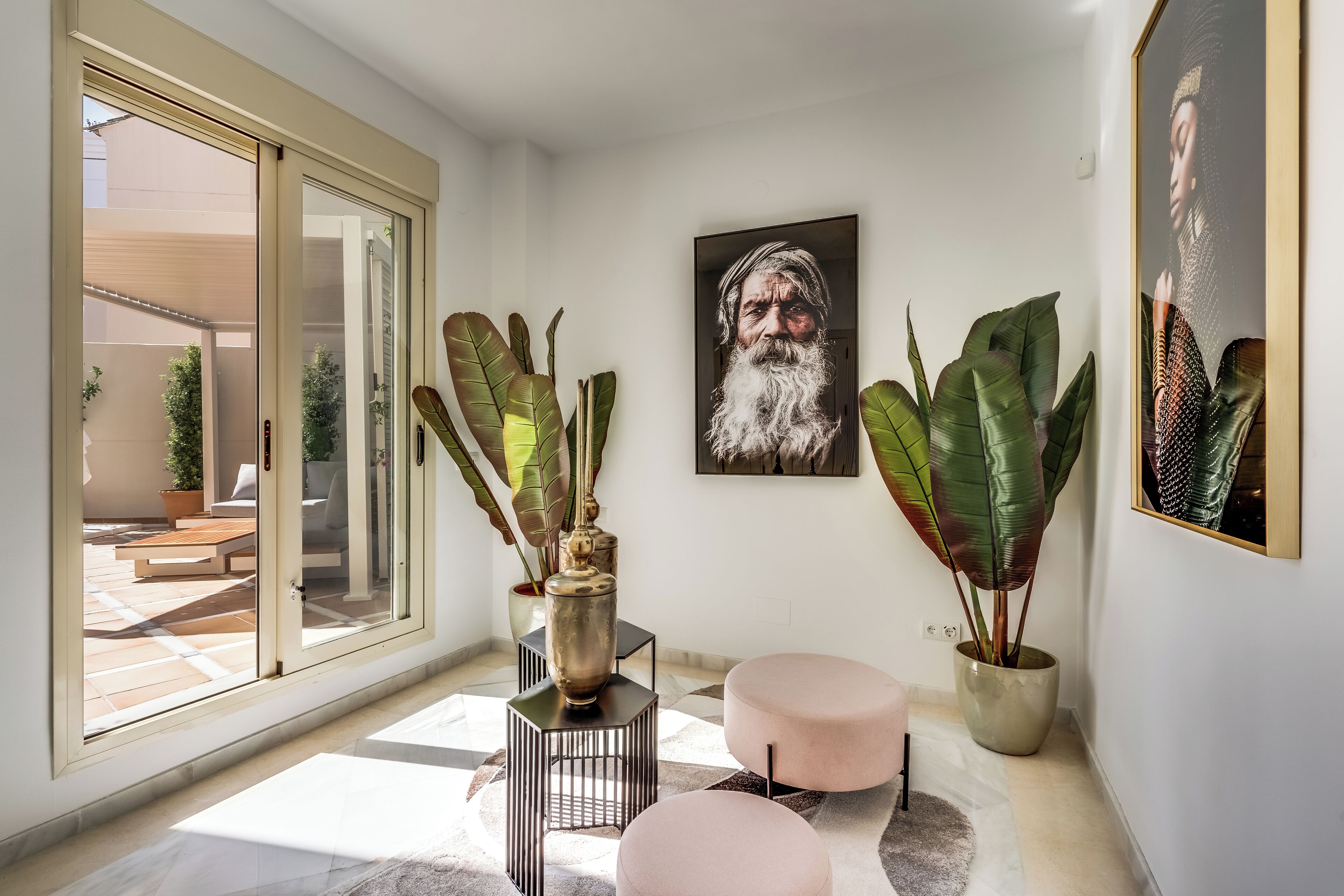 Un intérieur moderne avec une grande porte-fenêtre coulissante donnant sur un patio extérieur, une grande plante en pot, un portrait encadré et un petit pouf rond.