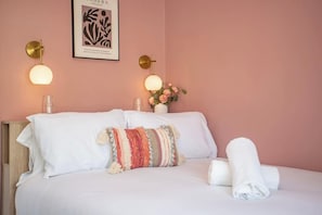 Una camera d'albergo con un letto, cuscini, una lampada a parete, un'opera d'arte incorniciata e un vaso con fiori.
