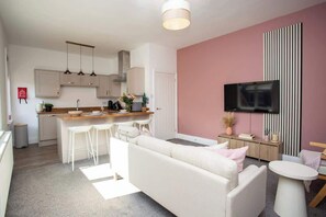 A modern kitchen with pendant lights, a dining area with white bar stools, a sofa, and a TV mounted on a pink wall.