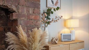 A brick wall, a white vase with dried plants, a lamp, and a small white radio on a wooden shelf.
