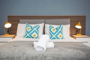 Ein ordentlich bezogenes Bett mit einem aufgerollten Handtuch, zwei Nachttischlampen und gemusterten Kissen.