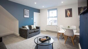 Una sala de estar moderna con un sofá gris, una mesa de centro de vidrio y una zona de comedor con sillas blancas.