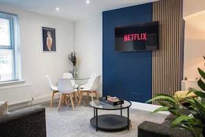 Ein modernes Wohnzimmer mit Netflix-Fernseher, einem Glastisch und einem Essbereich mit weißen Stühlen.