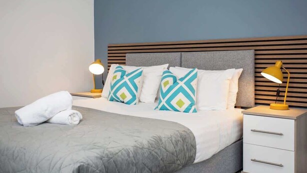 Una cama bien hecha con colcha gris, dos almohadas con diseño geométrico, una mesita de noche blanca con cajones y dos lámparas de noche.