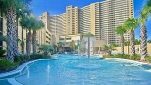A swimming pool with a water fountain, surrounded by palm trees and a high-rise hotel building in the background.