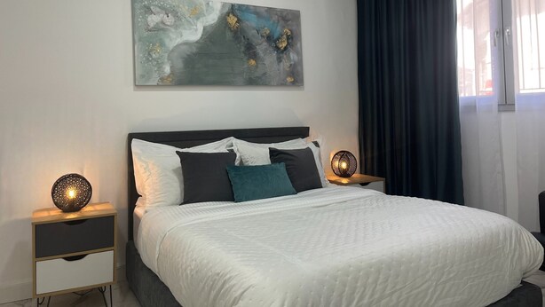 Un dormitorio con una cama, dos mesitas de noche, aire acondicionado, una obra de arte en la pared y una ventana con cortinas.