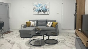 Una sala de estar moderna con un sofá seccional gris, dos mesas redondas, un cuadro de un paisaje urbano y un mueble bajo para televisión de madera.