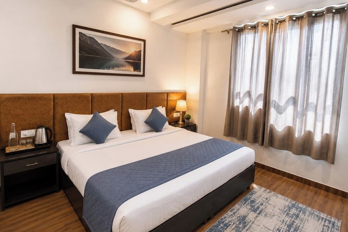 A hotel room with a bed, nightstand, lamp, and a painting of a mountain landscape.