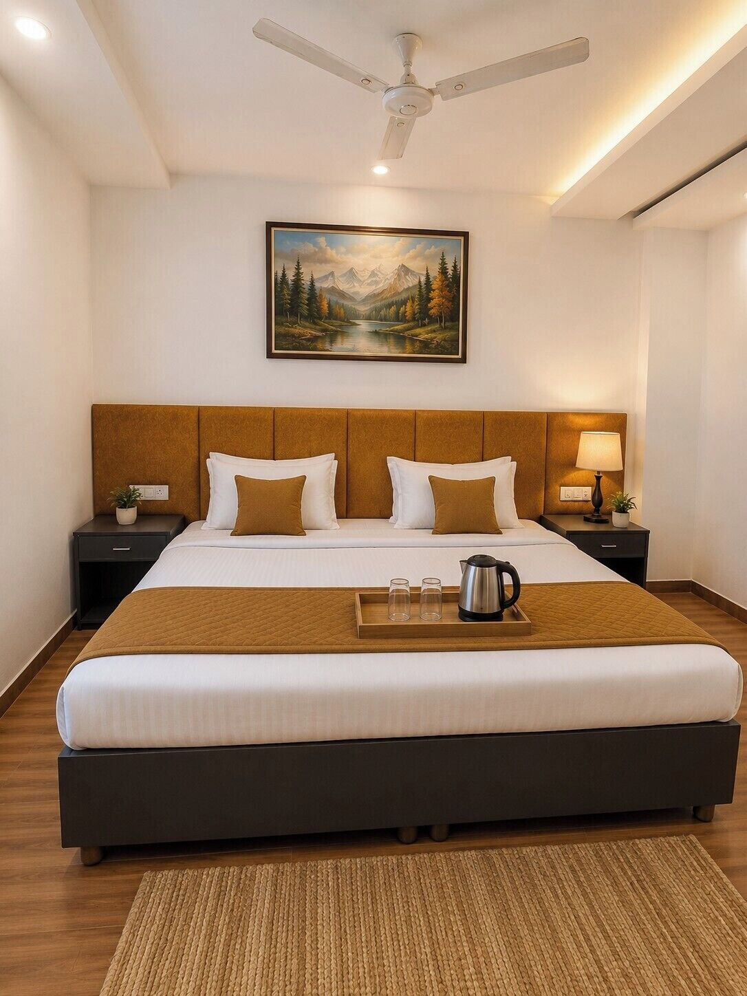 A hotel room with a large bed, a ceiling fan, a painting of a mountain landscape, bedside lamps, and a tray with glasses and a kettle.