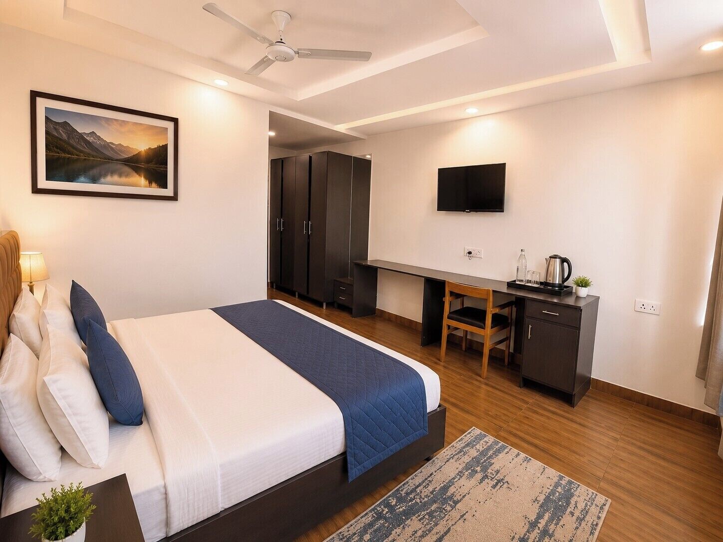 A modern hotel room with a bed, desk, wardrobe, TV, and a painting of a mountain landscape.