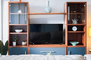 Un salon moderne avec un téléviseur à écran plat, des étagères en bois ornées d’objets décoratifs et un lit au premier plan.