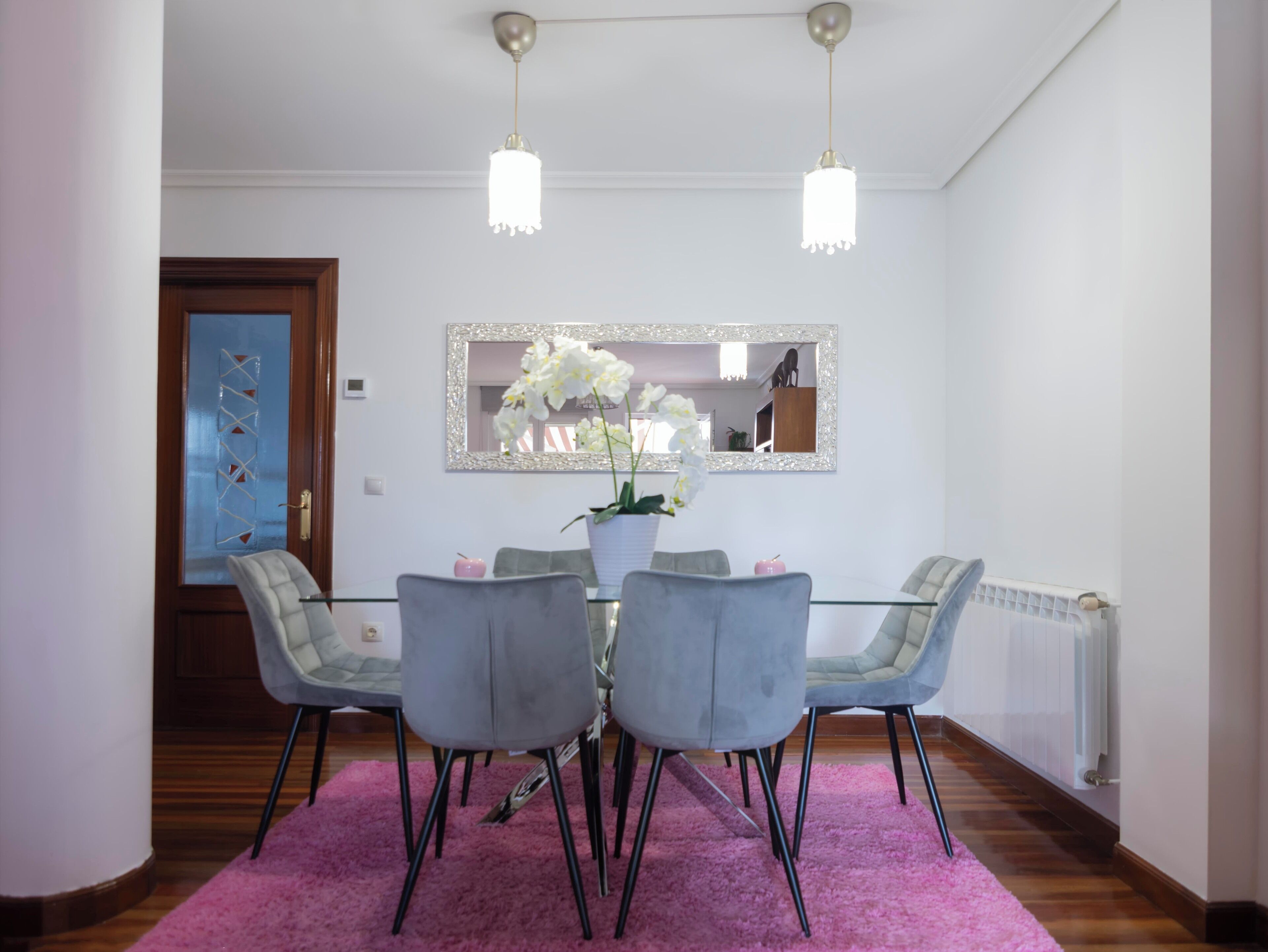 Spisestue med et glasbord, fire grå stole, en pink måtte og to hængende lamper.