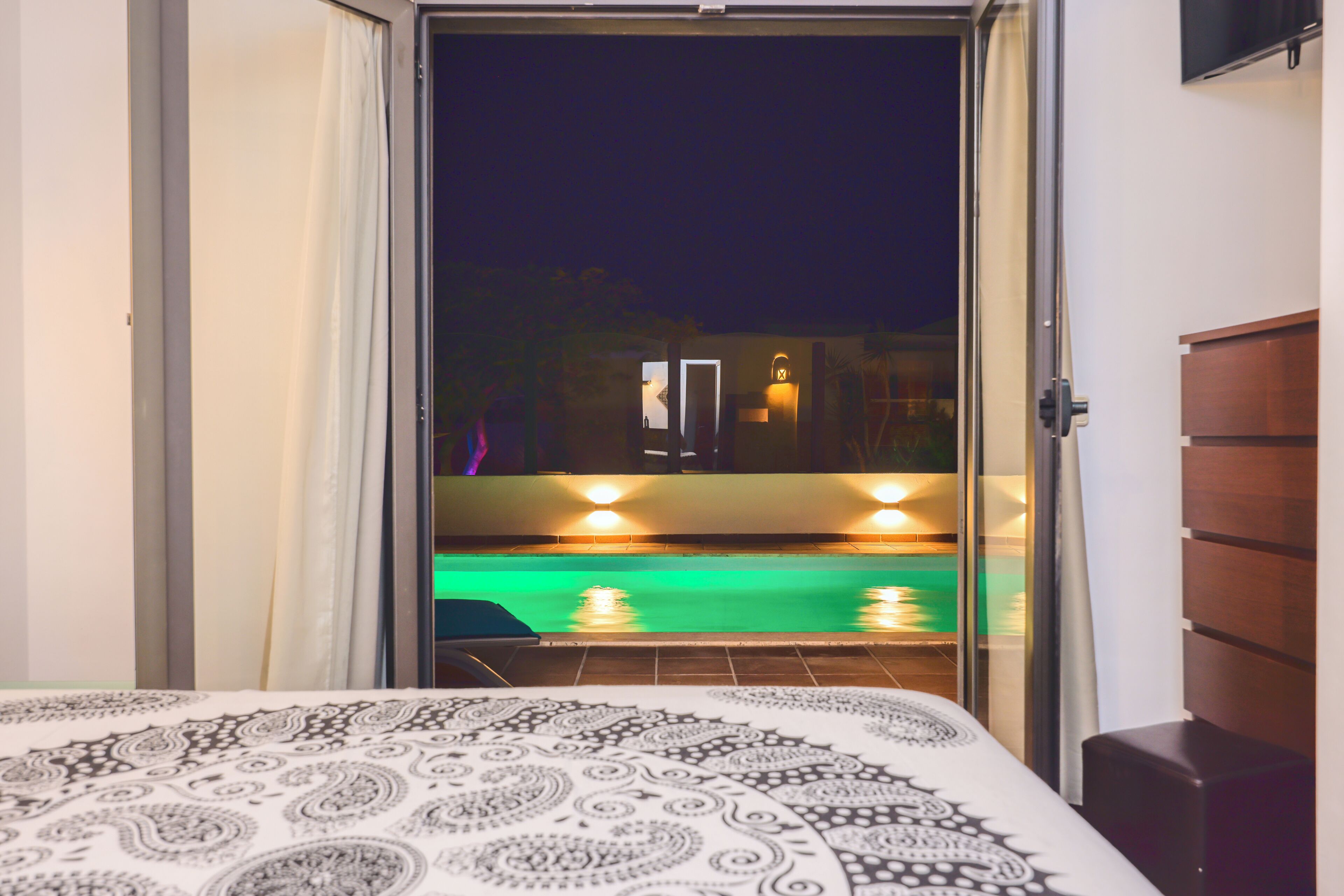 Une chambre avec un lit, une vue sur une piscine la nuit, et une porte donnant sur un espace extérieur.