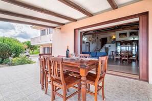 Uma área de refeições ao ar livre com mesa e cadeiras de madeira, um alpendre coberto e vista para o interior da casa.