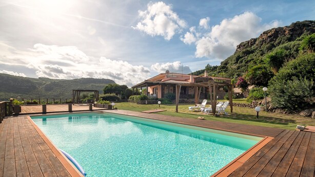 A wooden deck surrounding a clear blue pool with a house in the background. - Villa 'Patrizia' with Sea View, Wi-Fi and Air Conditioning (Castelsardo)