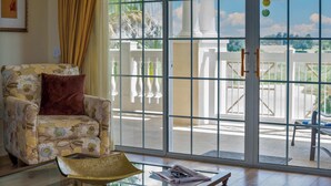 A room with a patterned armchair, a glass table with a magazine, and a view of a balcony with white railings and a palm tree. - Bright home near Disney w/ themed rooms & pool (Kissimmee)