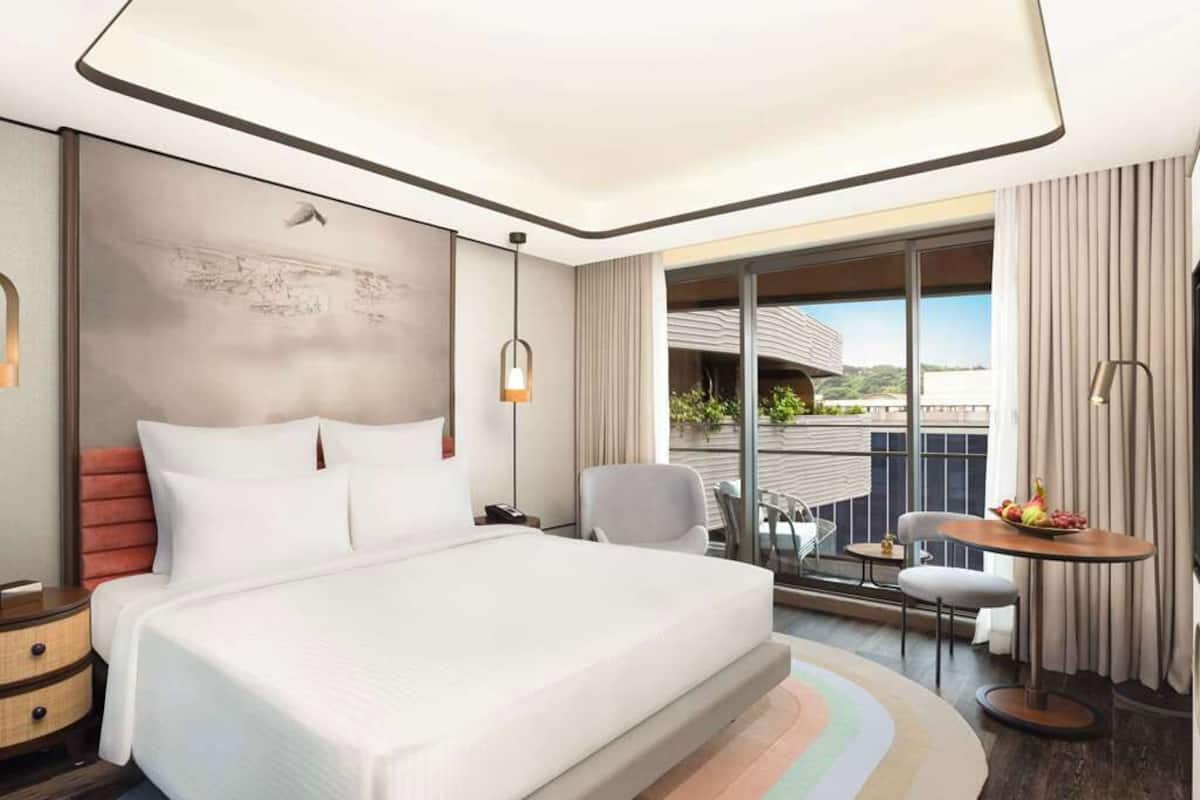 A modern hotel room with a large bed, a small table with fruit, a chair, a desk, and a balcony with a view.