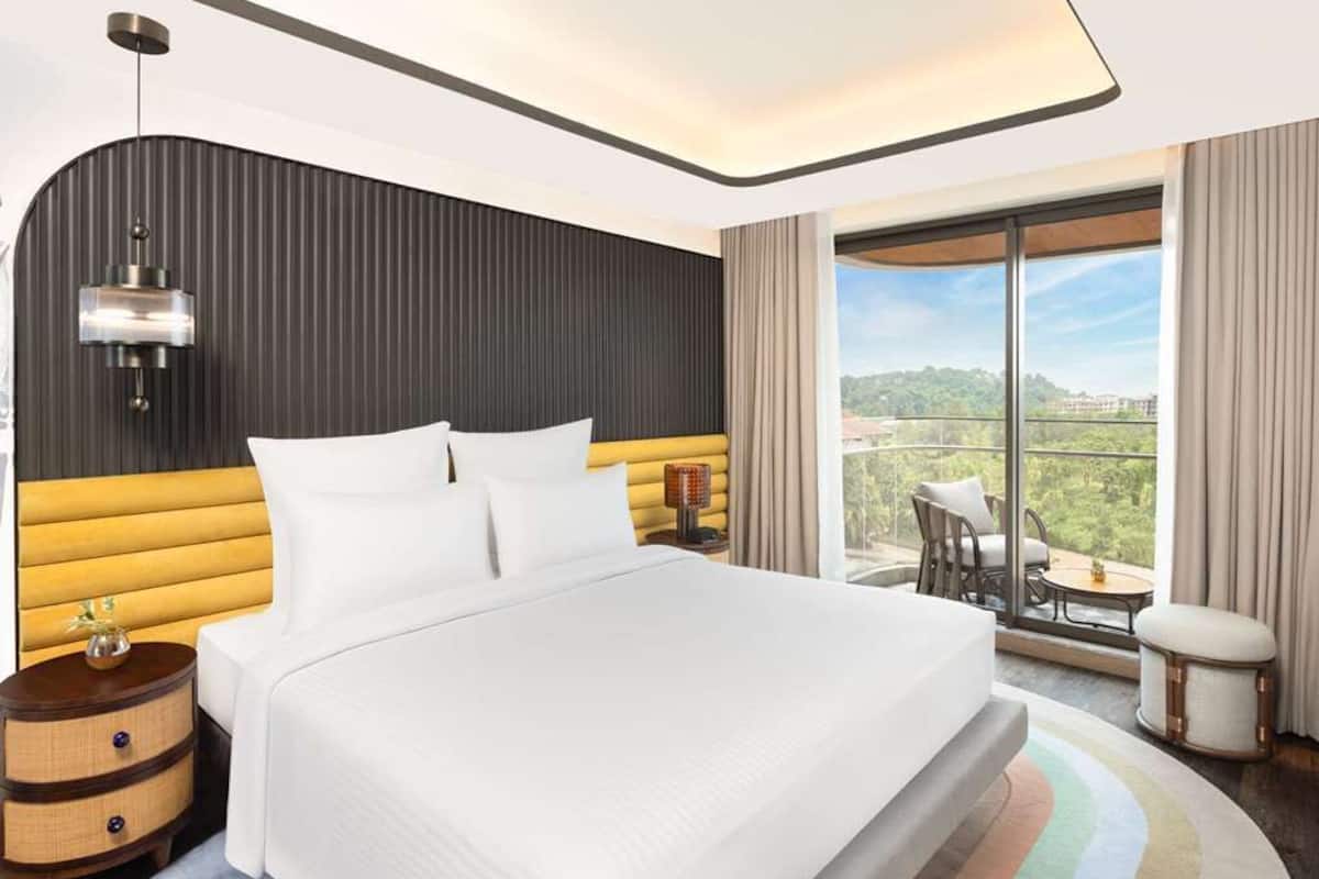 A modern hotel room with a large bed, a curved headboard, a nightstand, a lamp, and a view of the outdoors.