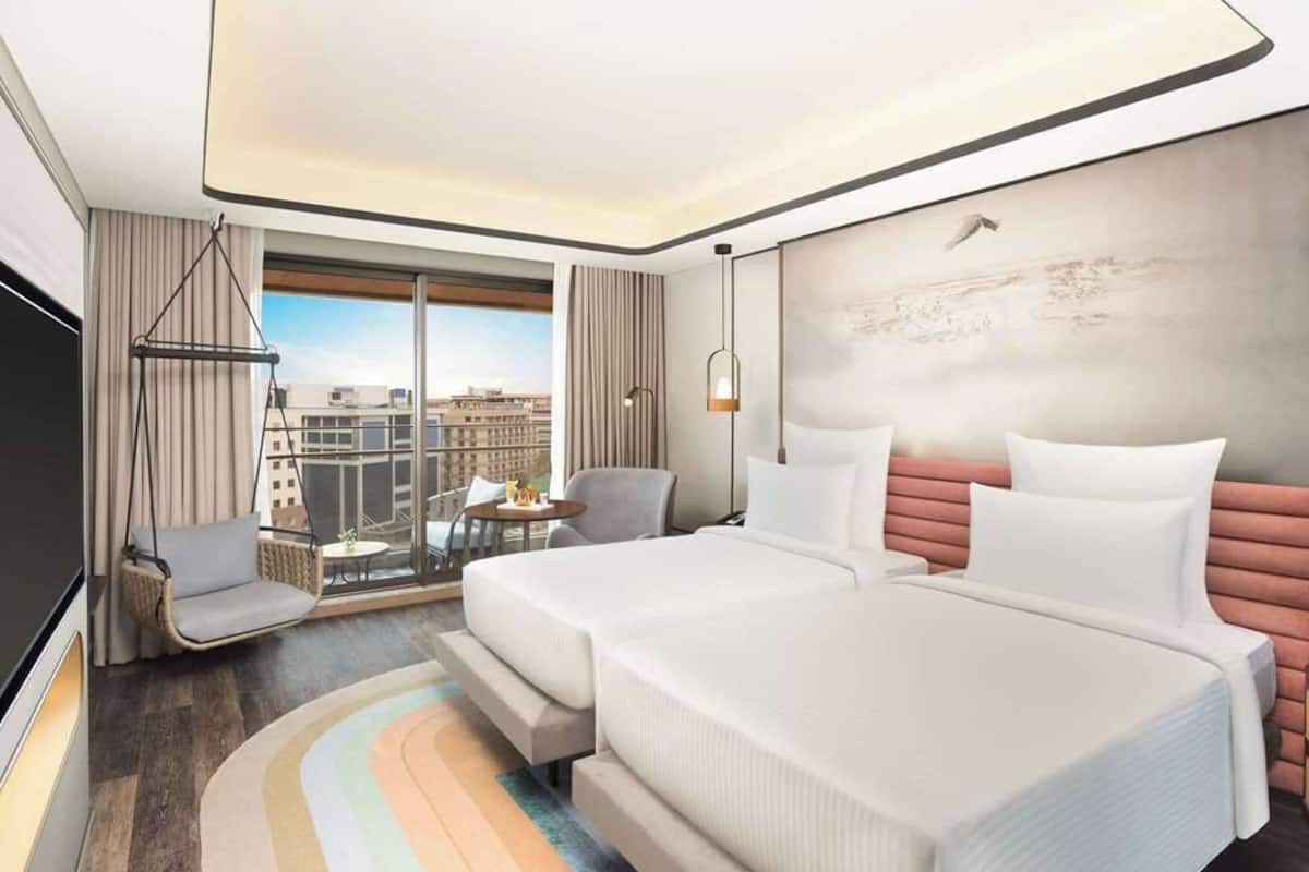 A modern hotel room with two beds, a sofa, a TV, a large window with a city view, and a hanging light fixture.
