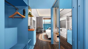 A modern hotel room with a large bed, a desk, and a TV. There is a bathroom with a shower and a sink. The room has a blue color scheme and a large window. - Novotel GOA Panjim (Panaji)