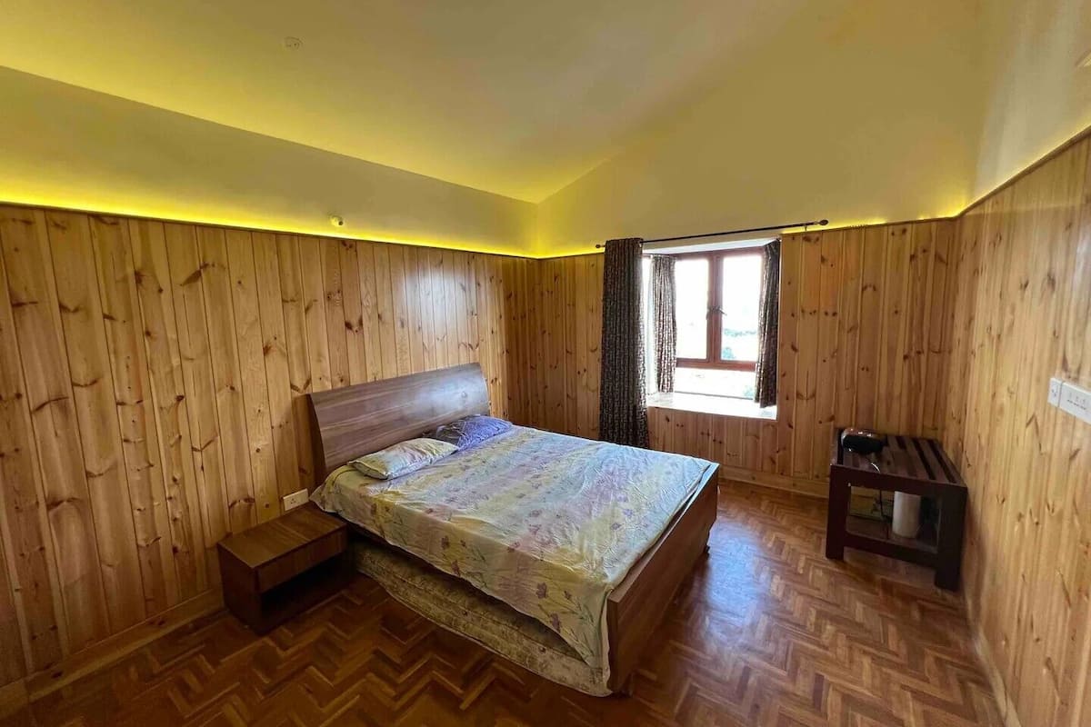 A bedroom with wooden panel walls, a bed with a floral bedspread, a small bedside table, and a window with curtains.