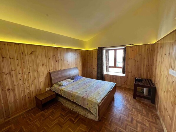 A bedroom with wooden panel walls, a bed with a floral bedspread, a small bedside table, and a window with curtains. - Sky Top Villa (Kodaikanal)