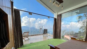 A room with a large sliding glass door leading to a balcony with a view of mountains. There is a bed, a chair, and a small table. - Sky Top Villa (Kodaikanal)