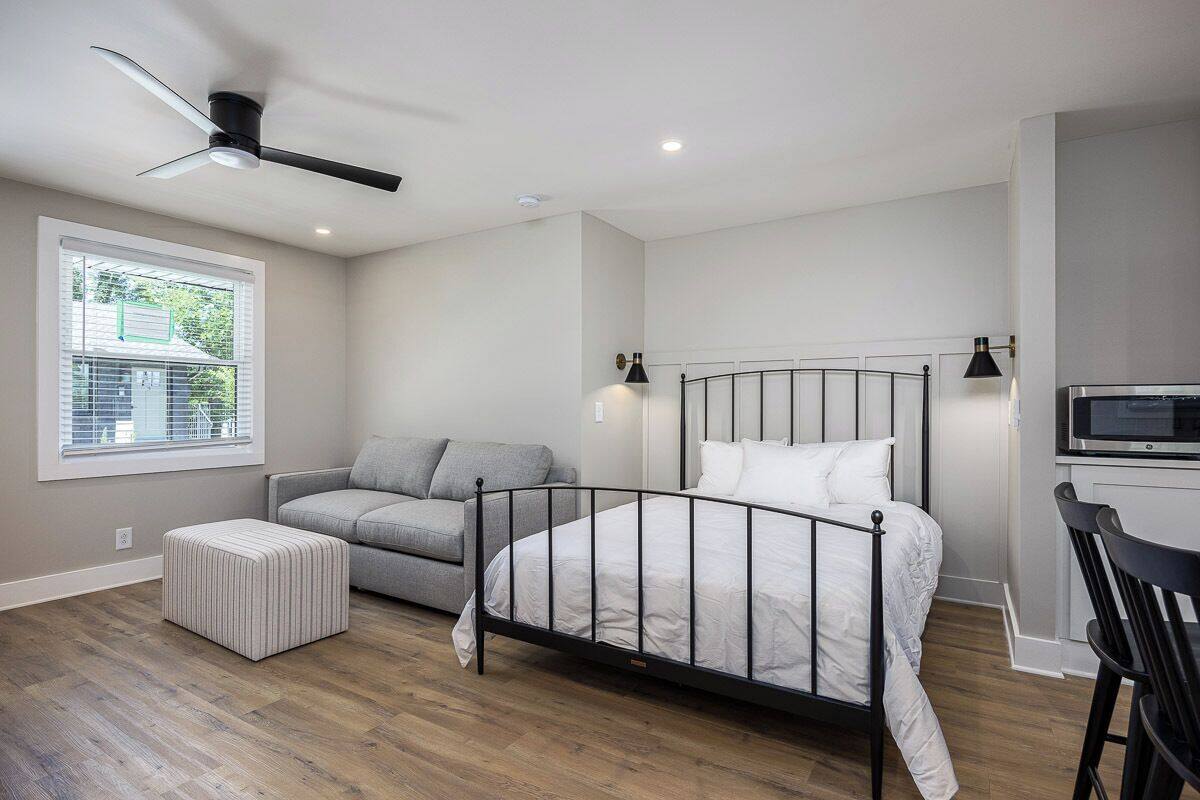 A bedroom with a bed, a grey sofa, a white ottoman, a ceiling fan, a TV, and a window with blinds.
