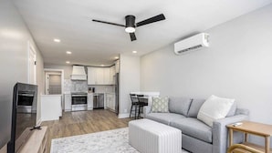 Een moderne woonkamer met een grijze bank, een houten salontafel en een gestreept voetenbankje. Er staat een televisie op een dressoir, een plafondventilator en een airconditioning aan de muur.