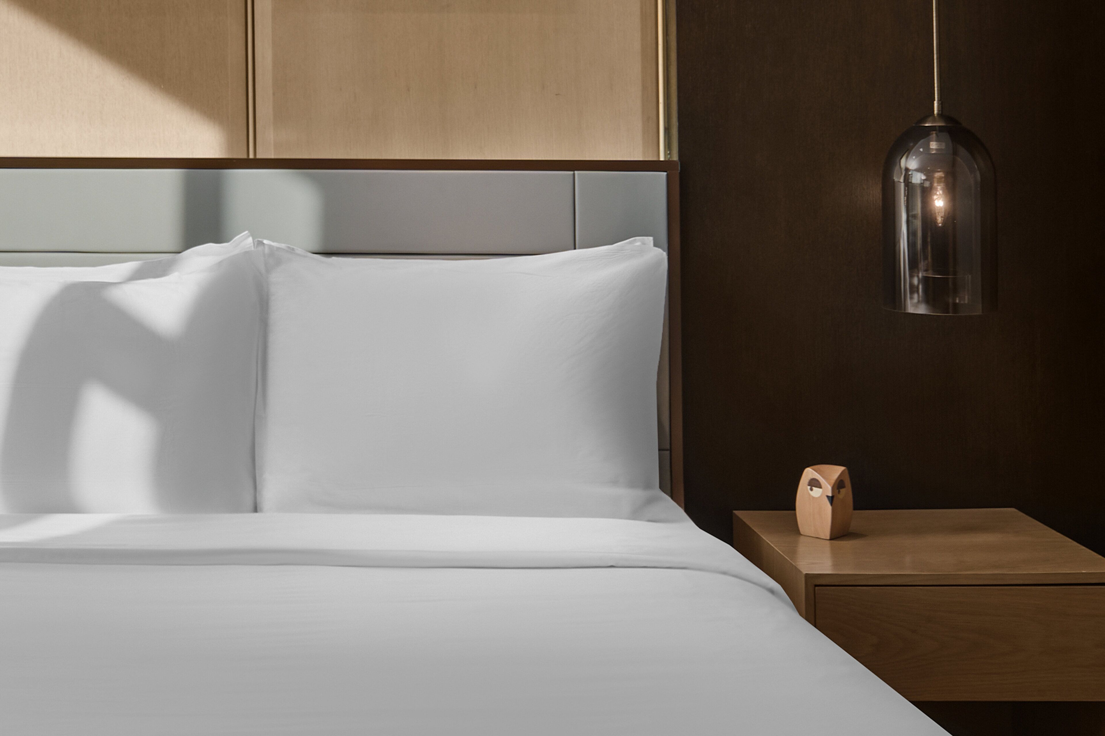 A neatly made bed with white linens, a wooden nightstand with a wooden clock, and a pendant light hanging above.