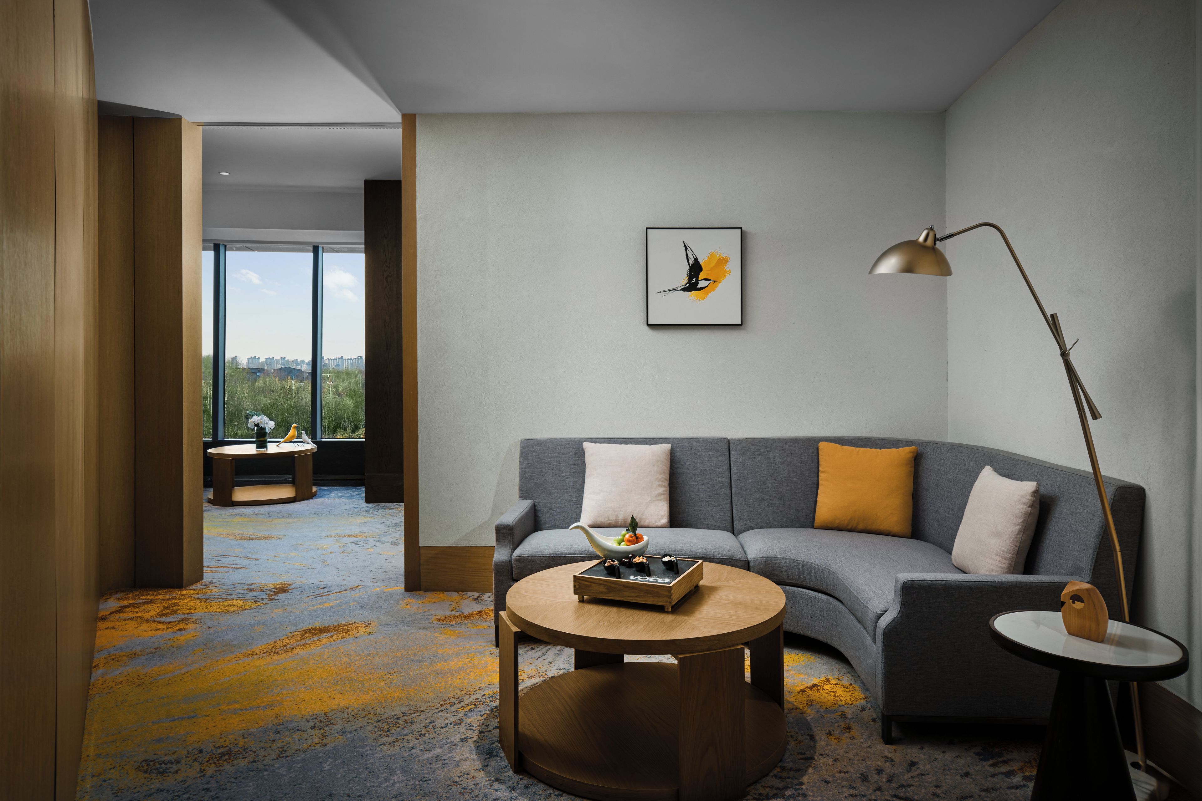 Una sala de estar moderna con un sofá gris, una mesa de centro redonda y una lámpara de pie. Hay una obra de arte enmarcada en la pared y una vista de la ciudad a través de ventanas amplias.